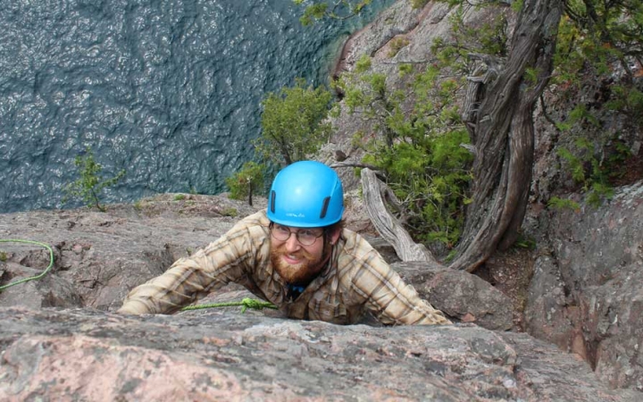 A person wearing safety gear and secured by ropes pauses their climb to look up at the camera. They appear to be high above a body of water. 